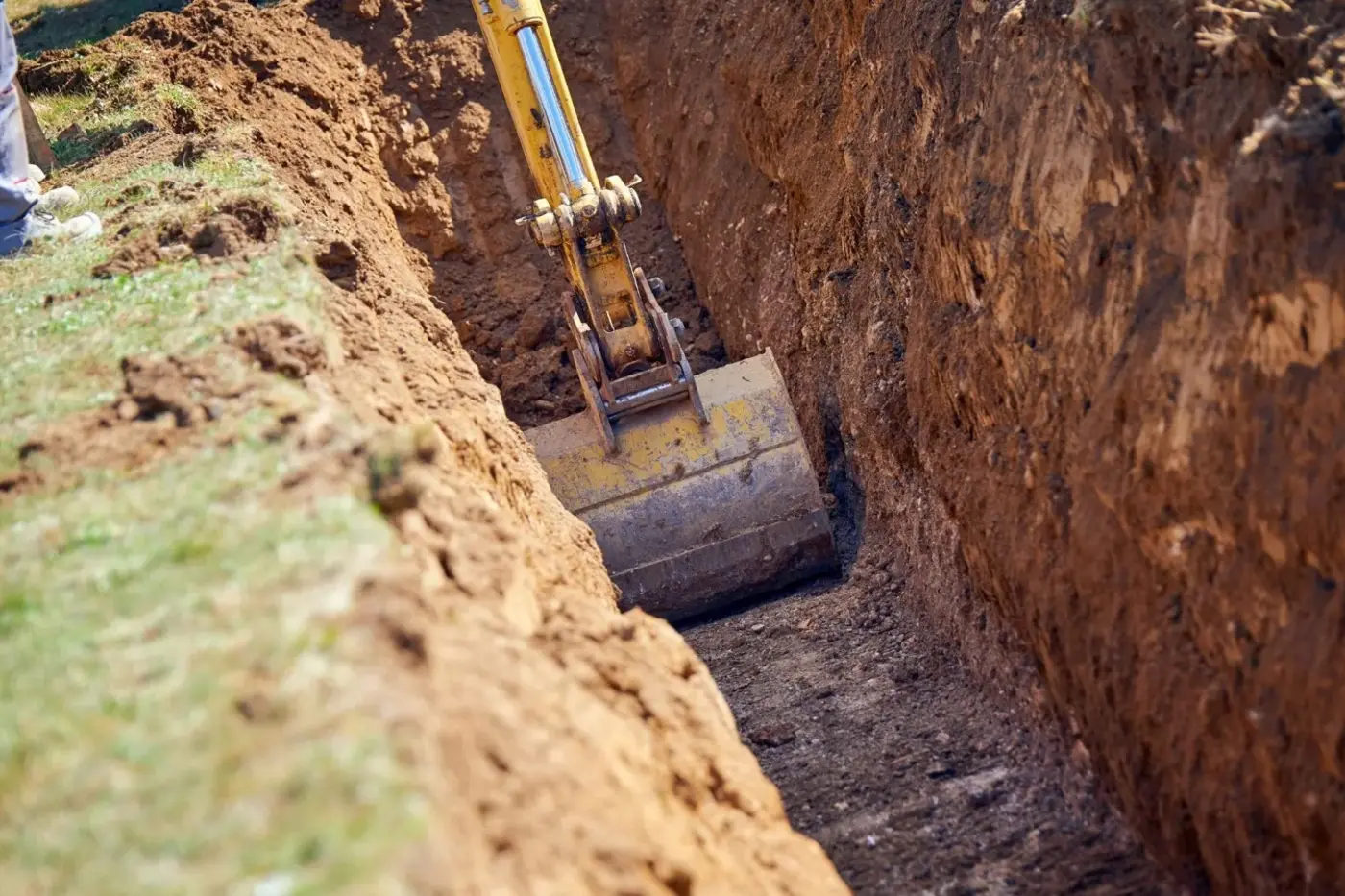 Excavator bucket trenching a narrow, deep cut through the soil beside a grass yard